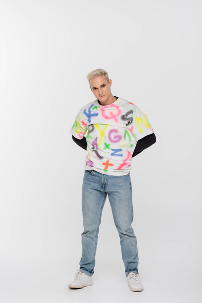 full length of gay man in jeans and t-shirt with alphabet print posing with hands behind back on grey background - Fotó, kép
