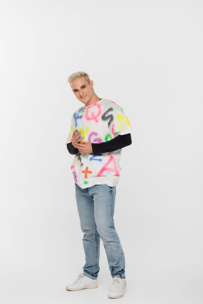 full length of blonde fashionable gay man smiling at camera on grey background - Photo, image