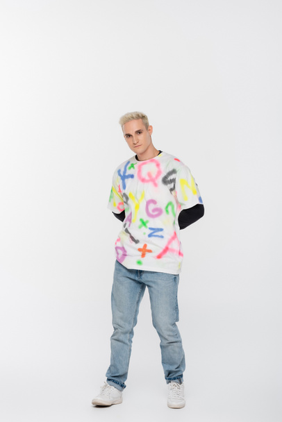 full length of young gay man in jeans and t-shirt with multicolored alphabet print on grey background - Fotó, kép