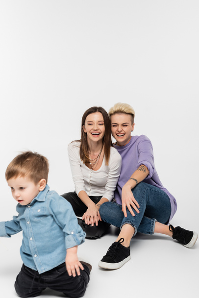 cheerful lesbian couple laughing near toddler son on grey background - Foto, afbeelding