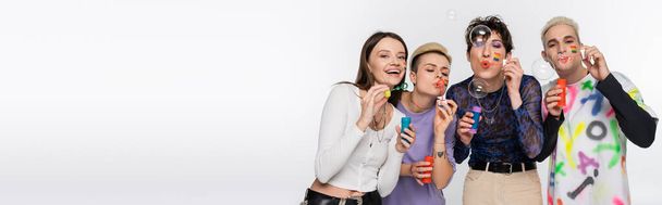 stylish people of different identity and with lgbtq flags on faces blowing soap bubbles isolated on grey, banner - Photo, Image