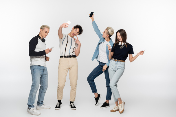 young man taking selfie and showing victory sign near friends with smartphones dancing on grey background - Zdjęcie, obraz