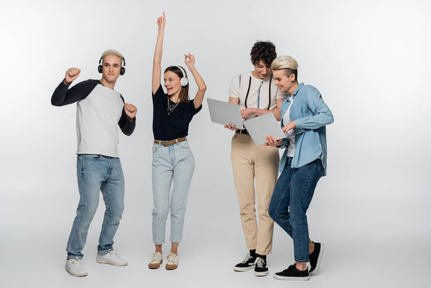 cheerful couple dancing in headphones near friends with laptops on grey background - Foto, afbeelding