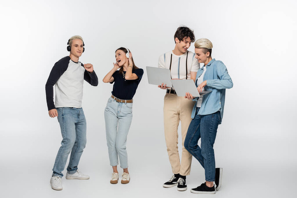 smiling couple using laptops near cheerful friends dancing in wireless headphones on grey background - Фото, изображение