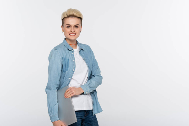 trendy woman in denim shirt looking at camera and holding laptop isolated on grey - Zdjęcie, obraz