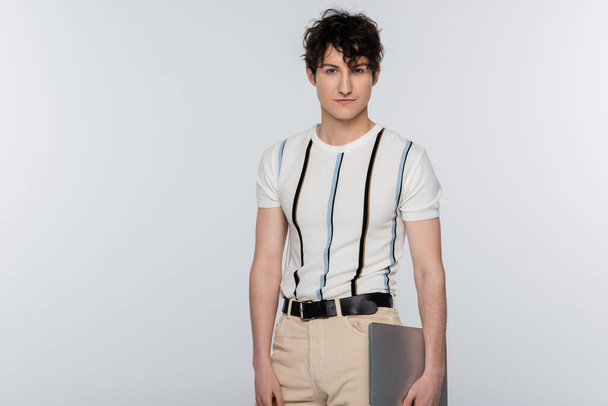 young man in striped t-shirt and beige trousers holding laptop isolated on grey - Fotoğraf, Görsel
