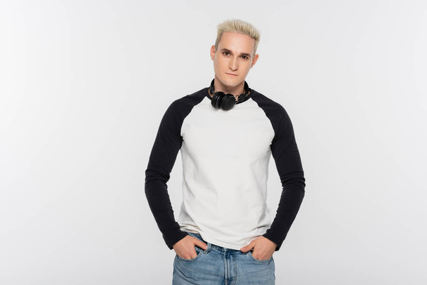 blonde man with headphones holding hands in pockets of jeans isolated on grey - Fotó, kép