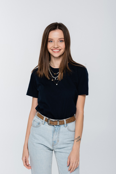 young brunette woman in black t-shirt and necklaces smiling at camera isolated on grey - Photo, image