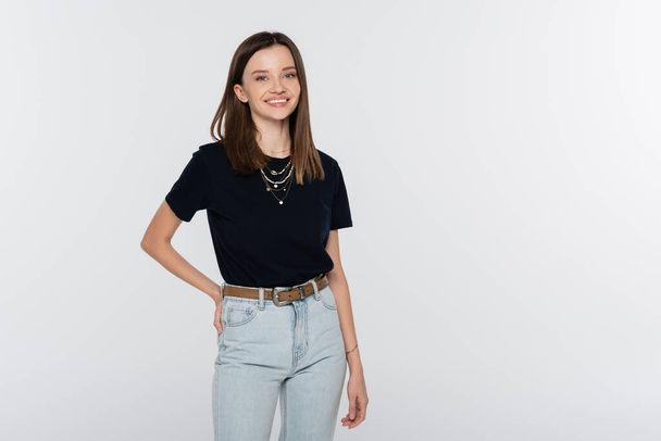 happy woman in black t-shirt and jeans posing with hand on hip isolated on grey - Fotó, kép