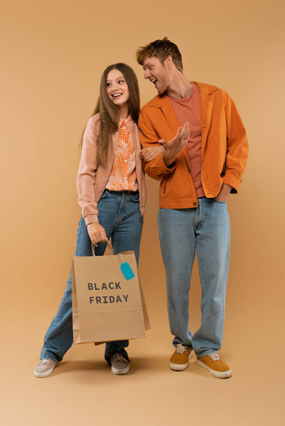 full length of young man talking near happy girlfriend holding shopping bag with black friday lettering on beige - Fotografie, Obrázek