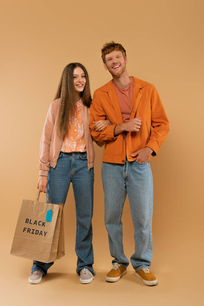 full length of young man posing near happy girlfriend holding paper bag with black friday lettering on beige - Photo, Image