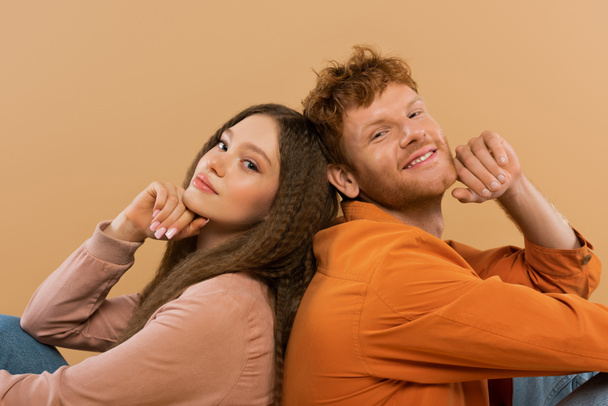 joyful young man with red hair sitting back to back with pretty girlfriend isolated on beige - 写真・画像