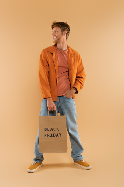positive young man with red hair holding shopping bag with black friday lettering and posing on beige - 写真・画像