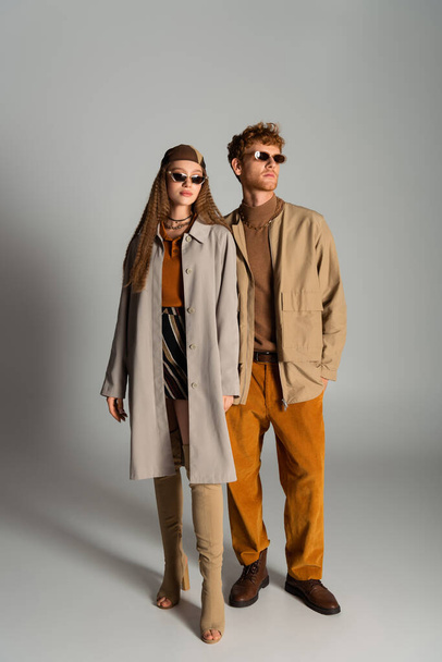 full length of young and stylish couple in autumnal outfits posing on grey - Foto, afbeelding