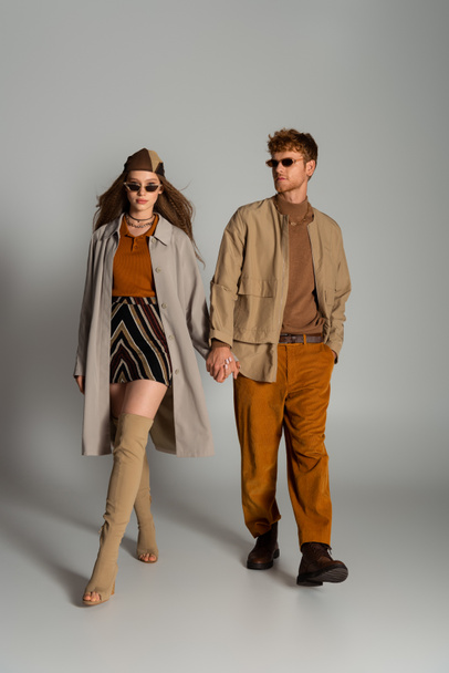 full length of young couple in stylish autumnal outfits and sunglasses holding hands while walking on grey - Foto, imagen