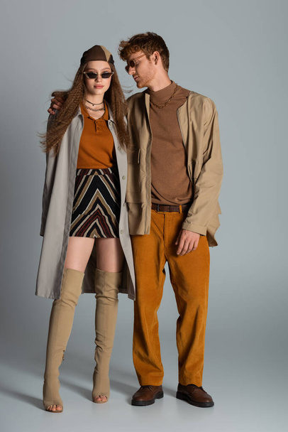 full length of young stylish couple in autumnal outfits and trendy sunglasses standing on grey - Photo, Image