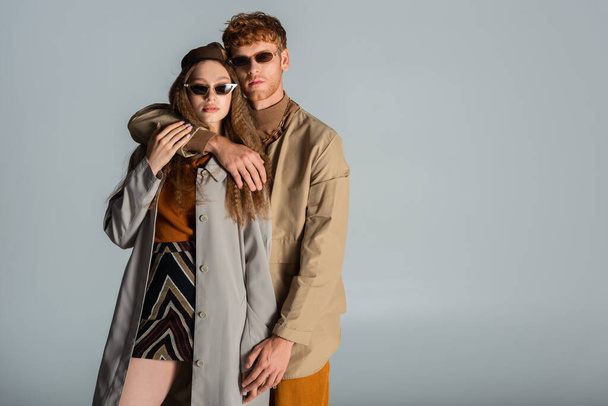 redhead man in sunglasses hugging stylish girlfriend in skirt and trench coat isolated on grey - Photo, image