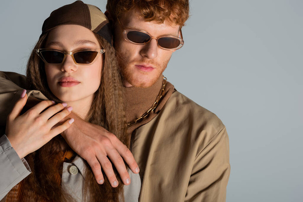 young redhead man in sunglasses hugging stylish girl in headscarf isolated on grey - Foto, afbeelding