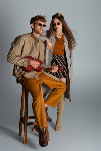 full length of redhead young man playing ukulele and sitting on high chair near girlfriend on grey - Photo, image