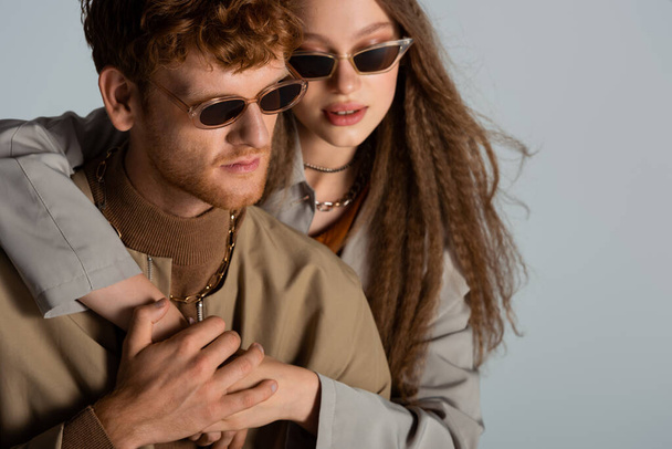 stylish teenage girl in sunglasses hugging redhead boyfriend isolated on grey - Φωτογραφία, εικόνα