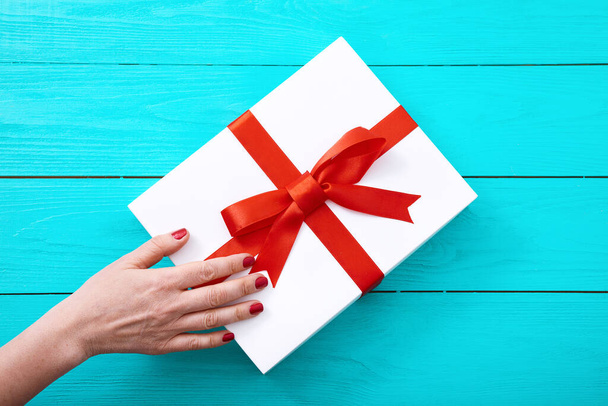 Female hand with red nails on gift box on blue wooden background. Top view - Foto, immagini