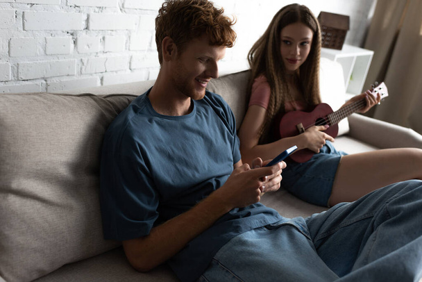 teenage girl playing ukulele and looking at happy boyfriend using smartphone while sitting on couch - Fotografie, Obrázek