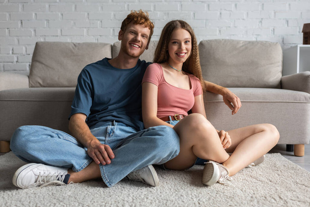 щаслива молода пара посміхається, сидячи біля дивана в сучасній вітальні
 - Фото, зображення