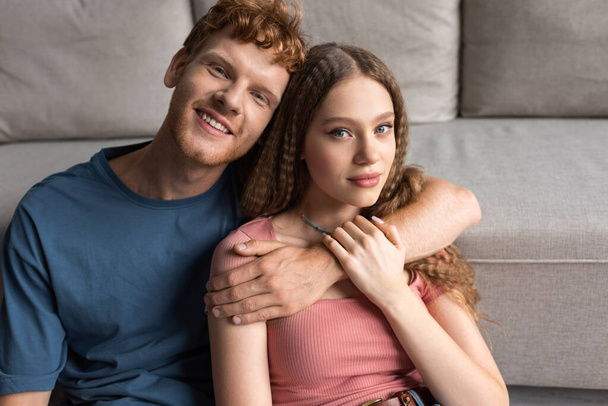 redhead young man smiling and hugging pretty girlfriend in living room  - Фото, изображение