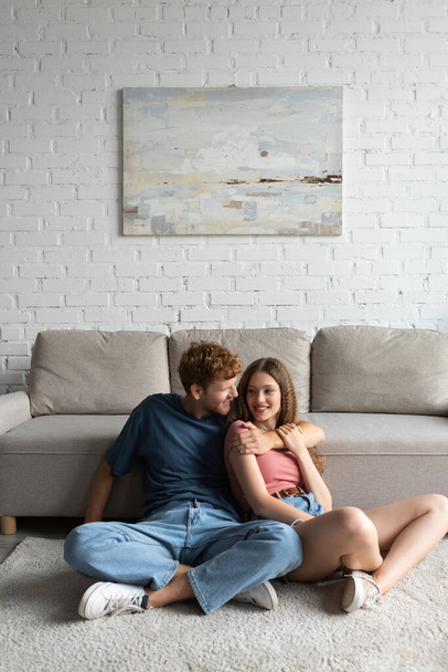 redhead and happy young man hugging and looking at cheerful girlfriend in modern living room  - Foto, Imagen