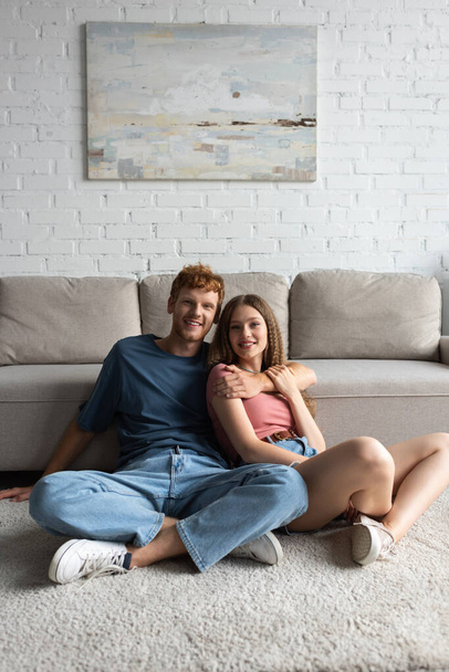 redhead and happy young man hugging pretty girlfriend in living room  - Foto, afbeelding