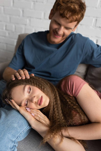 redhead and happy young man stroking hair of sleepy girlfriend in living room  - Foto, Bild