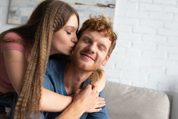 pretty teen girl hugging and kissing cheek of redhead boyfriend in living room - Fotó, kép