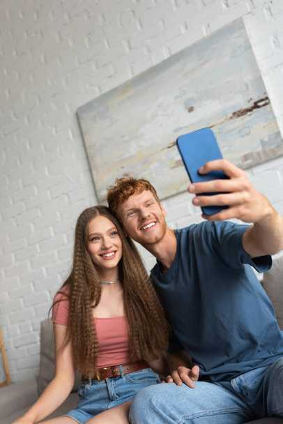 young and happy man taking selfie with cheerful girlfriend in living room - Foto, Imagen