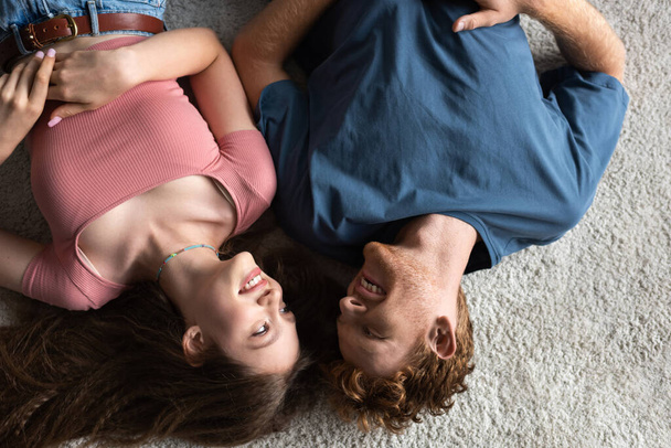 top view of cheerful young couple smiling and lying together on carpet in living room - Fotoğraf, Görsel