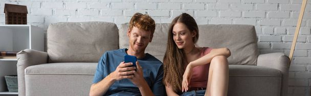 young redhead man using smartphone while sitting near smiling girlfriend and couch in living room, banner - Φωτογραφία, εικόνα