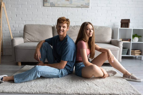 full length of happy young couple sitting on carpet and looking at camera in living room  - Valokuva, kuva