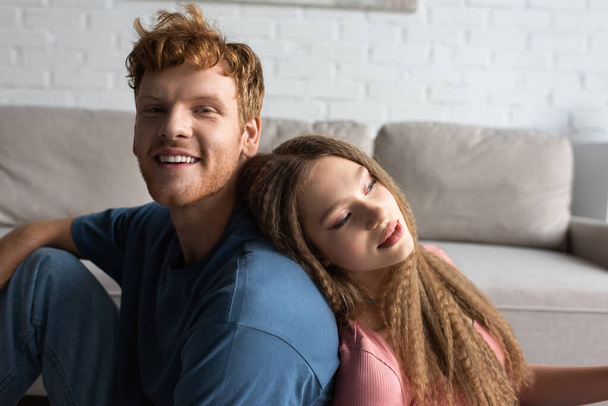 pretty teenage girl leaning on back of positive boyfriend with red hair in living room  - Фото, изображение
