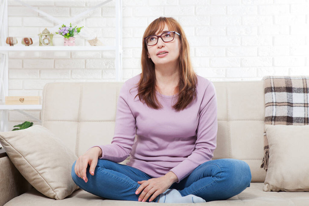 dreaming Middle-aged brunette woman with glasses and smile sitting on sofa at home background. Menopause and depression. Mock up and copy space - Photo, Image
