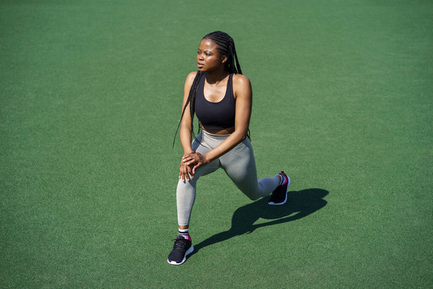 Młoda Afroamerykanka cieszy się utrzymaniem zdrowego trybu życia podczas gorącego letniego dnia. Sportowa dama z długimi warkoczami efektywnie ćwiczy mięśnie ciała na zielonej nawierzchni stadionu miejskiego - Zdjęcie, obraz