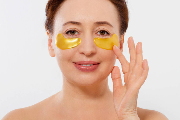 Beauty portrait of middle age woman with wrinkles and a gold patch under eye isolated on white background. Collagen mask and spa concept. Copy space. Summer skin care - Photo, Image