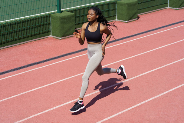 Atleta africana segura corriendo en el hipódromo. Deporte deportivo negro mujer entrenamiento trote en el estadio prepararse para el evento de la competencia, maratón o triatlón. Mezcla raza chica corredor practicando al aire libre - Foto, imagen