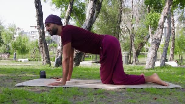 An arabian man stands on all fours with his legs crossed on a yoga mat in a park. Mid shot - Video