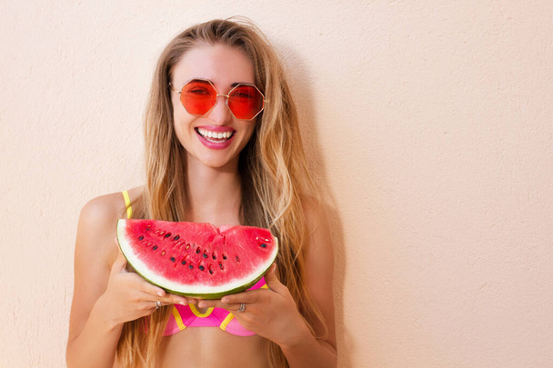 Summer taste. Cheerful young happy woman in swimsuit and sunglasses holding slice of watermelone standing outdoors - Фото, изображение