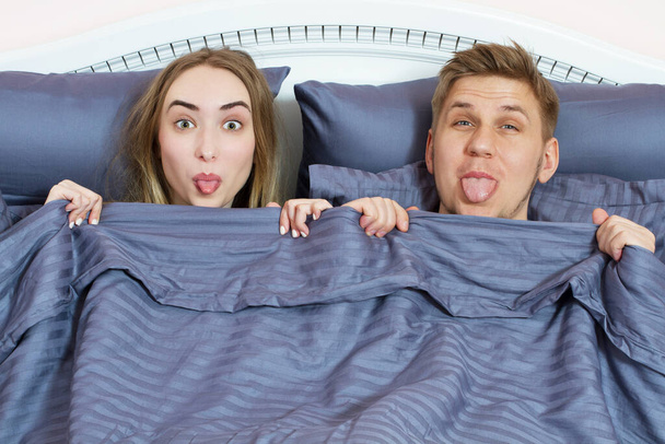 dorosłych pozytywna para w łóżku pod kołdrą pokazująca język i patrząca w kamerę - Zdjęcie, obraz
