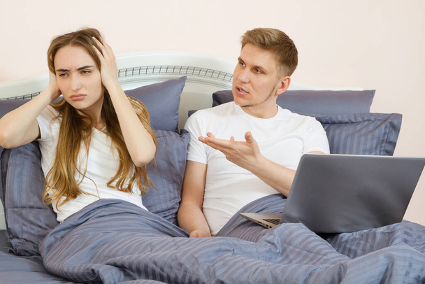 Στρεσαρισμένο νεαρό ζευγάρι συζητά κάτι στο κρεβάτι στο υπνοδωμάτιο με φορητό υπολογιστή, στρεσαρισμένο κορίτσι στο κρεβάτι - Φωτογραφία, εικόνα