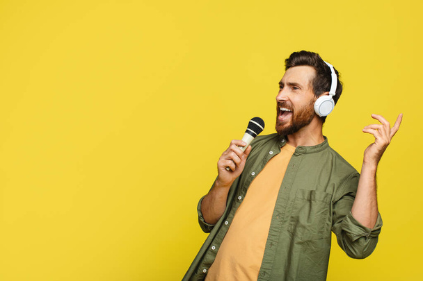 Радостный мужчина средних лет в наушниках, слушающий музыку и поющий песню в микрофон, мужчина в наушниках, наслаждающийся караоке на жёлтом фоне студии, копирующее пространство - Фото, изображение