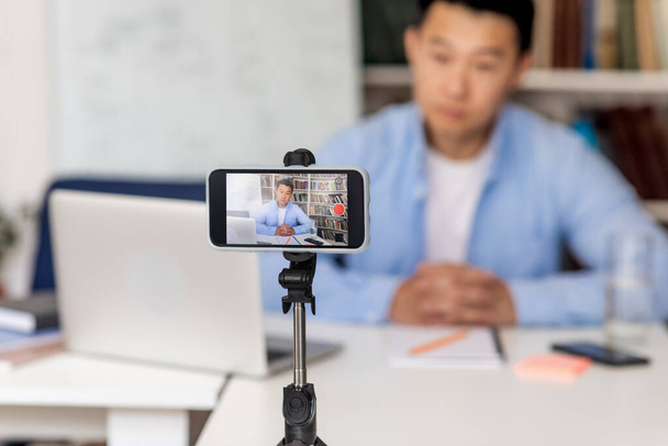 Электронное обучение. Серьезный китайский учитель мужского пола делает видео для образовательного блога через смартфон, сидя на рабочем месте в школе. Современное дистанционное образование. Избирательный подход к мобильным телефонам - Фото, изображение