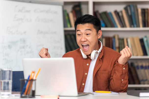 Emotional Asian Teacher Man Using Laptop Gesturing Yes In Joy Celebrating Educational Success Sitting At Workplace In Classroom. Successful E-Teaching Concept. Selective Focus - Photo, image