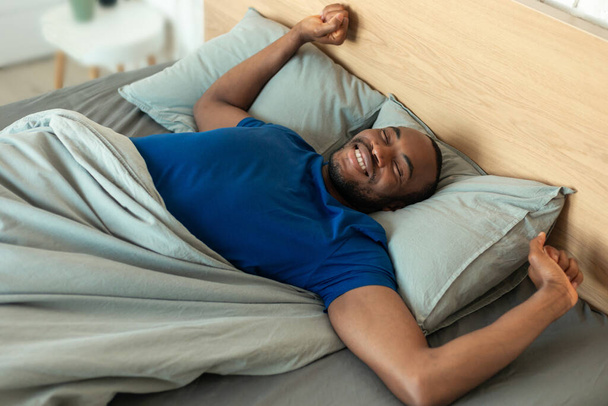 Happy Black Male Stretching Arms After Good Sleep Lying In Bed In Modern Bedroom Indoor. Wellslept Man Enjoying Lazy Weekend Morning. Healthy Sleeping Routine Concept. Above View - Photo, Image
