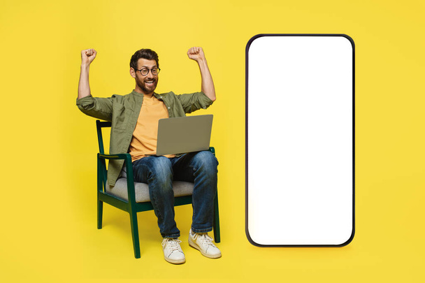 Excited caucasian man with laptop making YES gesture, sitting in chair near huge smartphone with mockup on screen, winning online lottery or casino bet, yellow studio background - Fotoğraf, Görsel